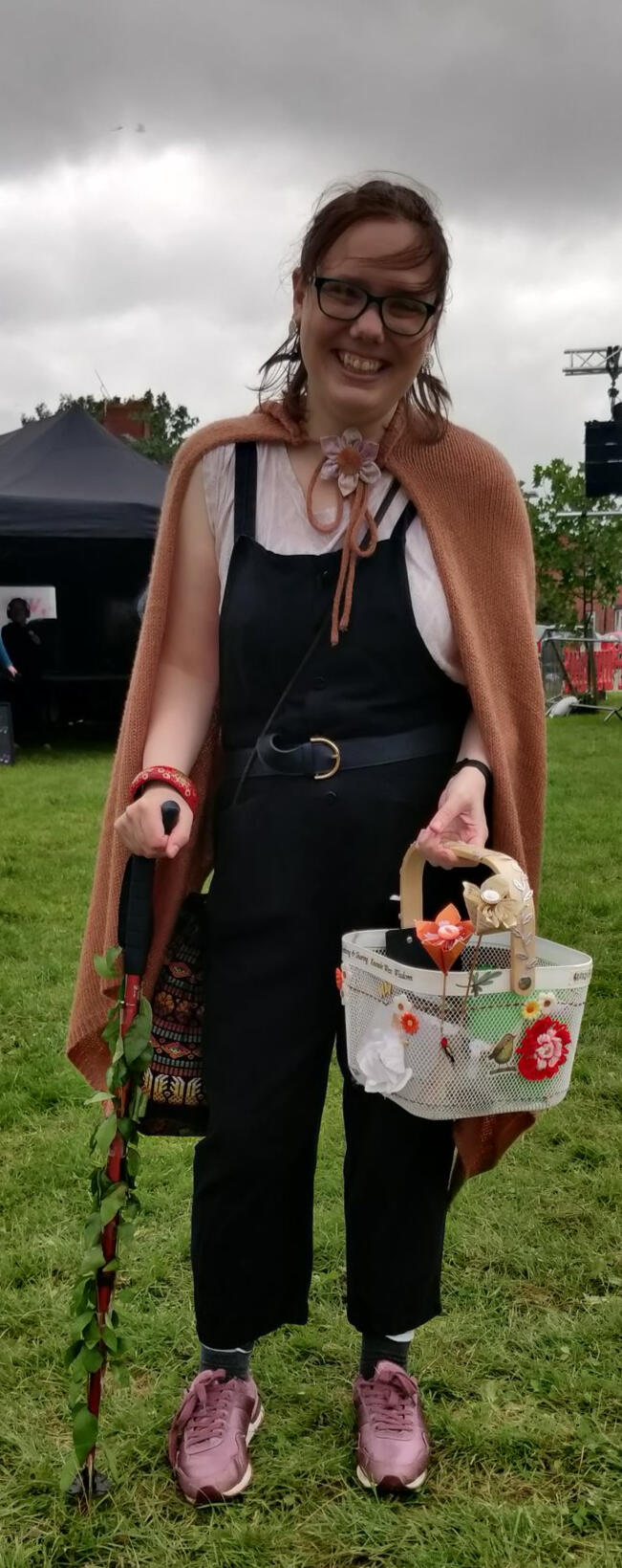 Me, a white woman with brown hair, outside on grass by a stage, dressed in knitted cape and dungarees, holding a decorated wisdom gathering basket and my walking stick, which is wrapped with leafy vines