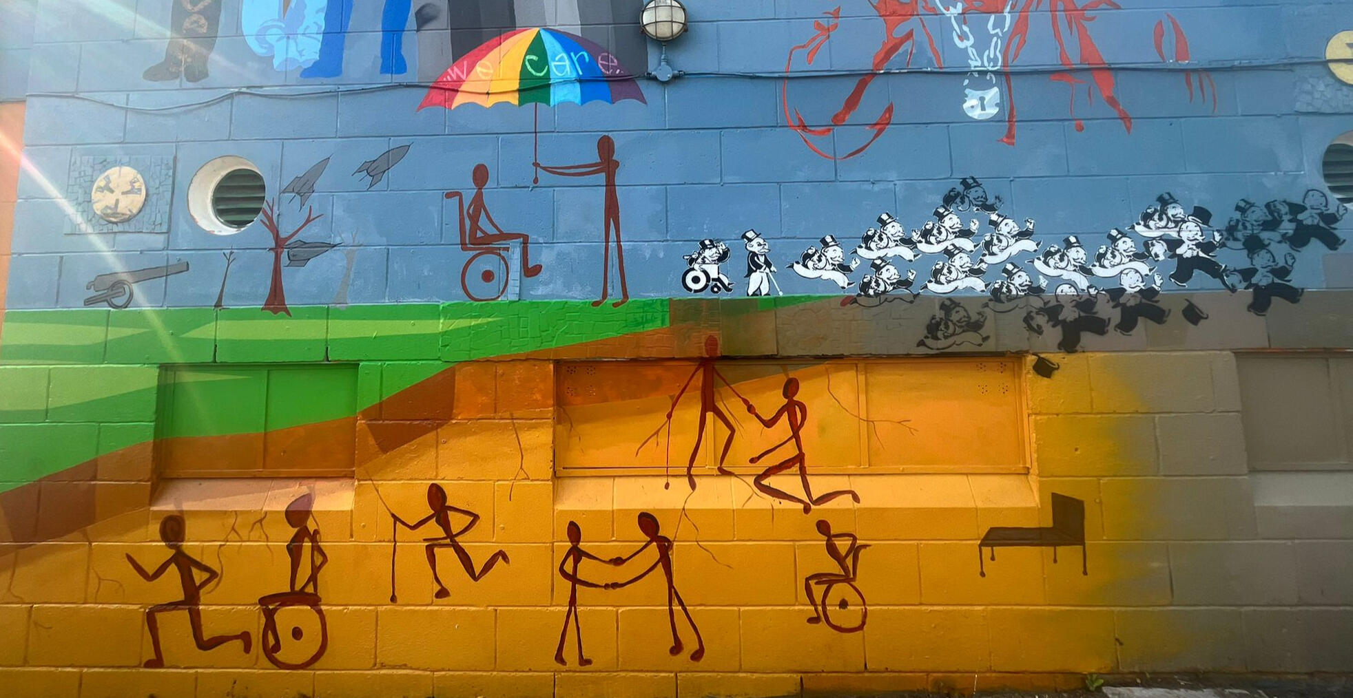 a colourful outdoor mural, showing cavemen style figures undergroup in wheelchairs, with sticks and holding hands in heart shapes, and helping each other, an empty bed, a canon shooting rockets past a wheelchair using caveman figure with another caveman fi